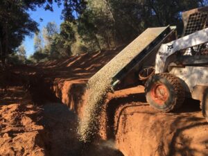 A compact skid steer equipped with a Skid Steer Side Dump Bucket deposits gravel into a trench in a wooded area, with trees in the background and a clear sky above.